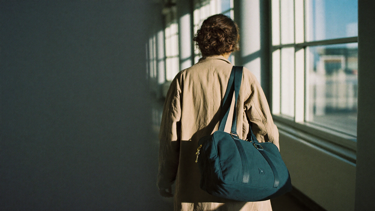Voyageuse de dos dans un couloir d'aeroport ensoleille, sac de voyage bleu canard a l'epaule